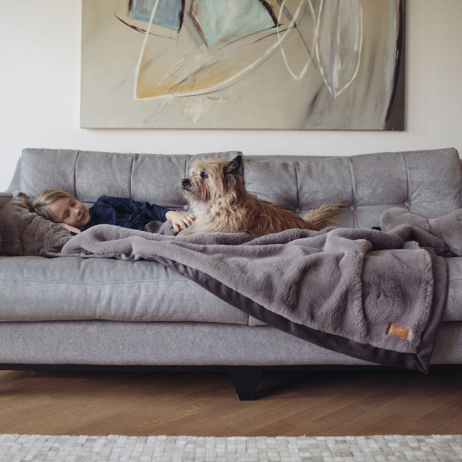 Mädchen liegt mit wuscheligem Terrier auf grauem Sofa unter der flauschigen grauen Hyggeplaid Hundedecke