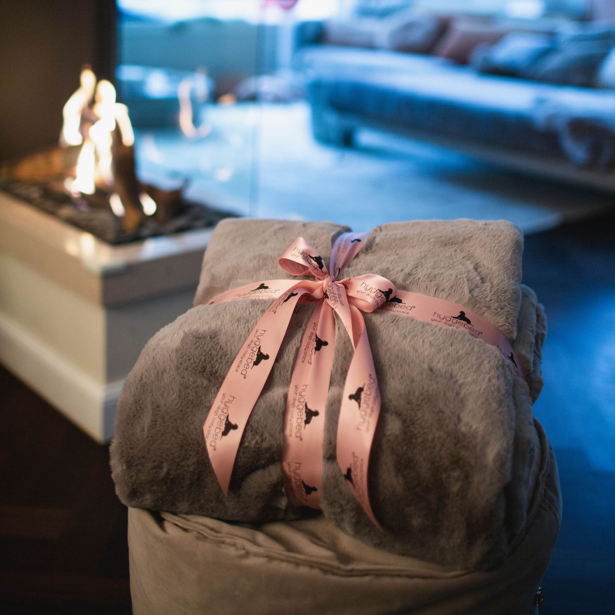 Zusammengerollte braune Hyggeblanket Kuscheldecke mit rosa Schleife neben einem brennenden Kamin