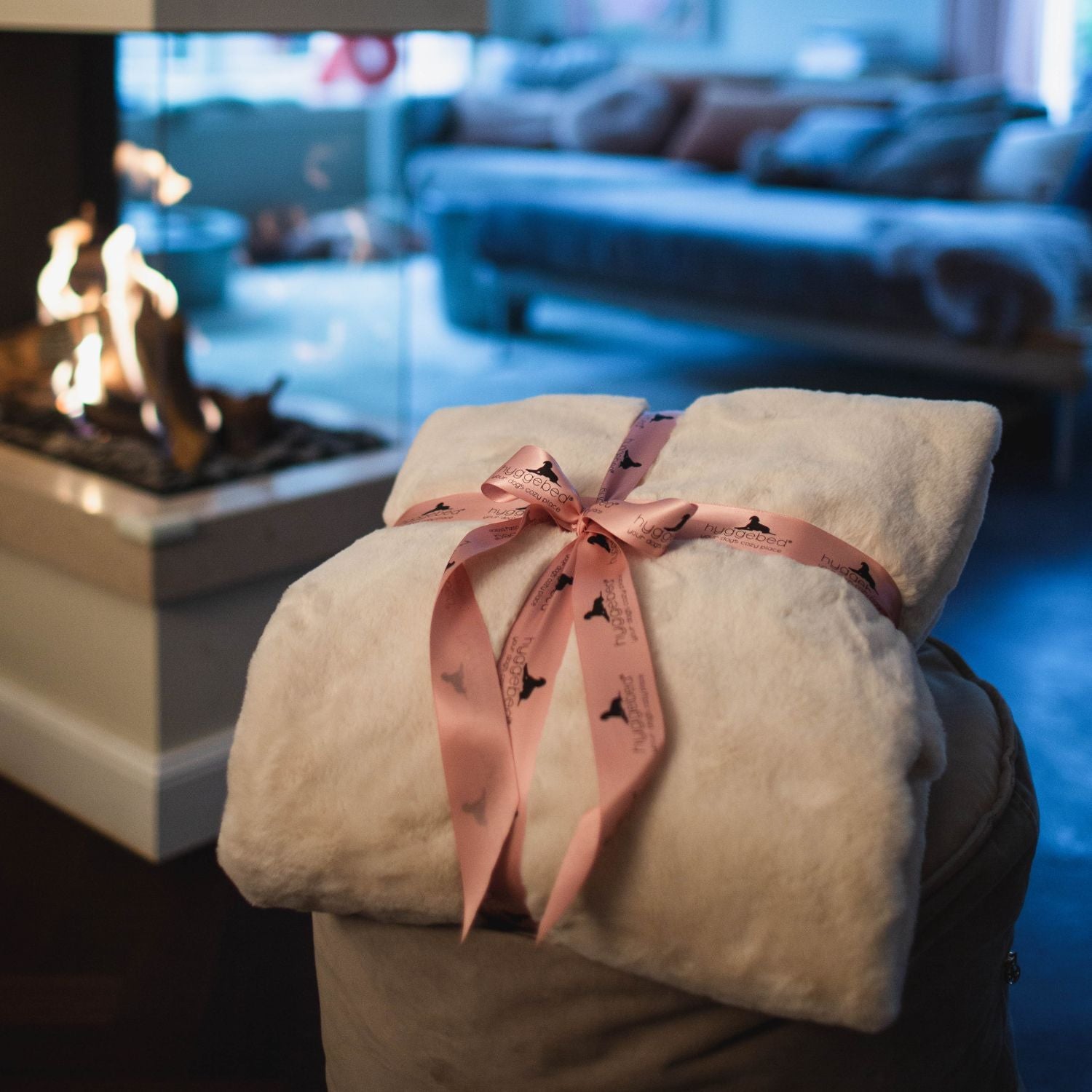 Zusammengerollte cremeweiße Hyggeblanket Decke Ivory mit rosa Schleife neben brennendem Kamin