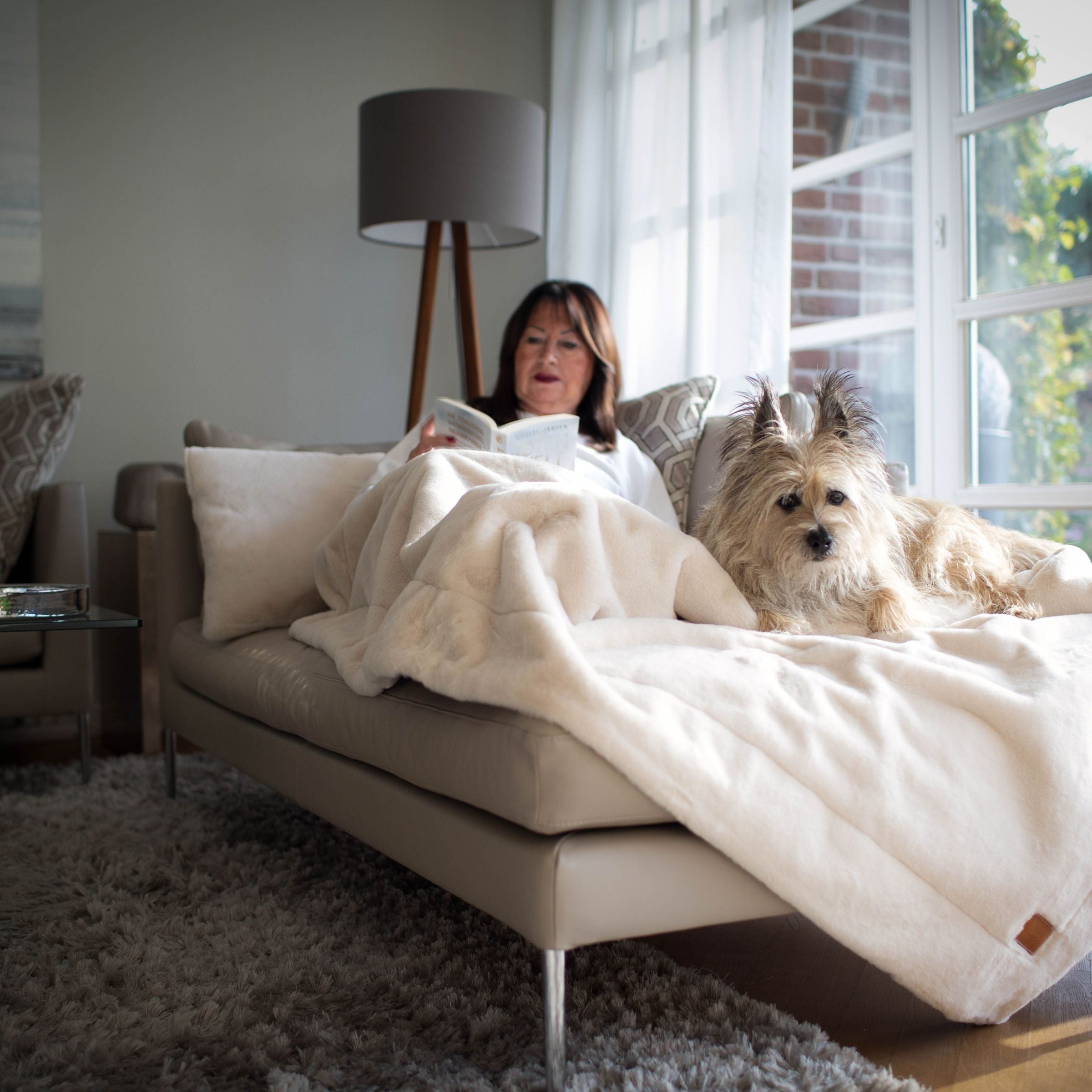 Frau liest ein Buch auf einer Chaiselongue unter der cremeweißen Hyggeblanket Decke neben einem aufmerksamen Hund