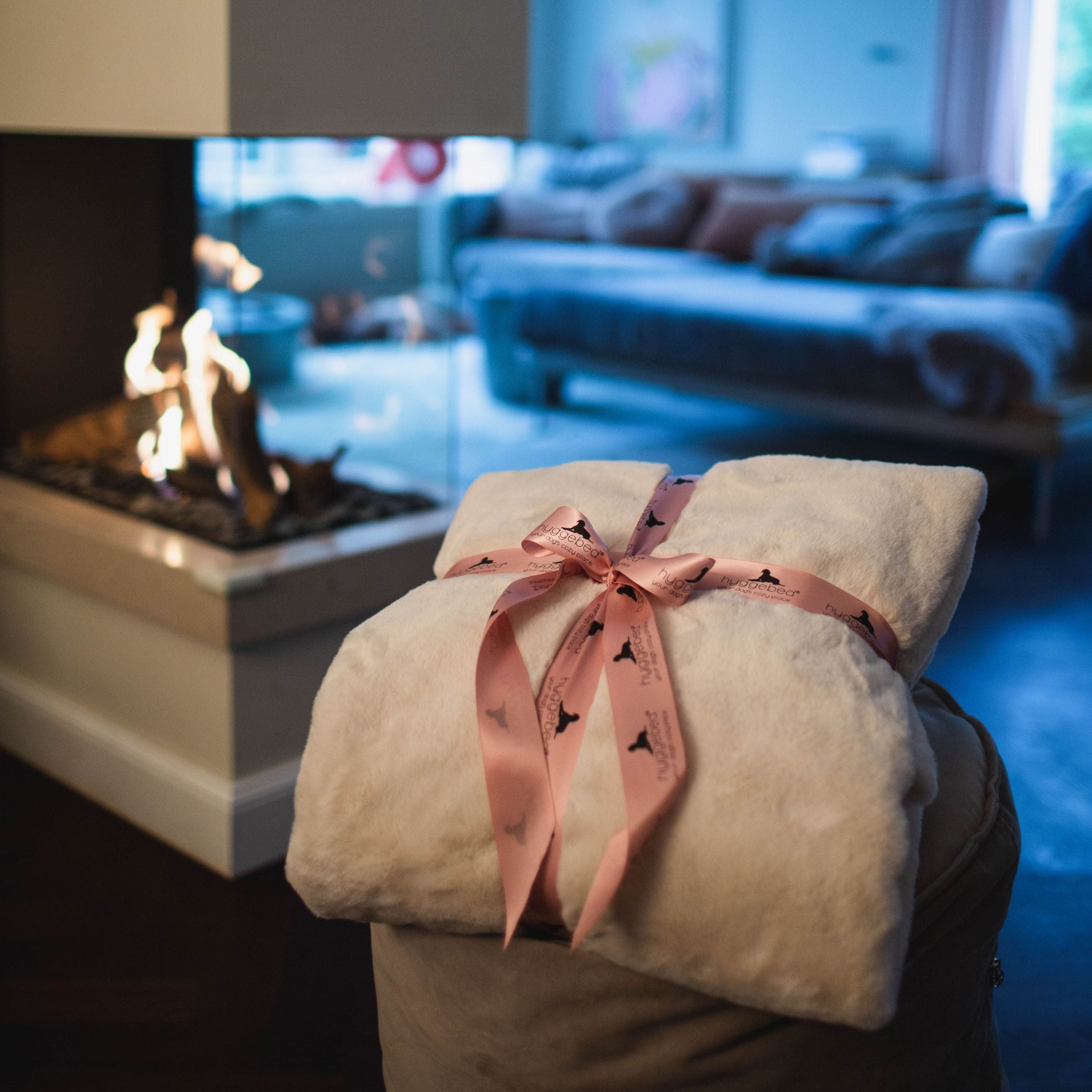 Zusammengerollte cremeweiße Hyggeblanket Kuscheldecke Ivory mit rosa Schleife neben einem brennenden Kamin