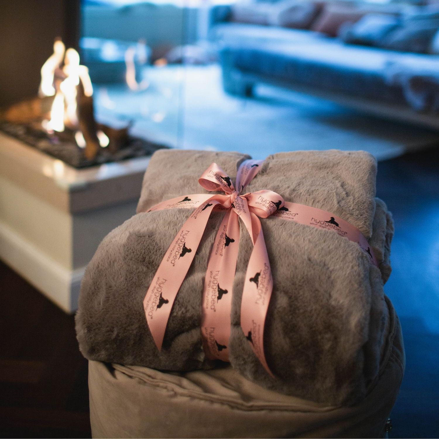 Zusammengerollte graue Hyggeblanket Decke Stone mit rosa Schleife neben brennendem Kamin
