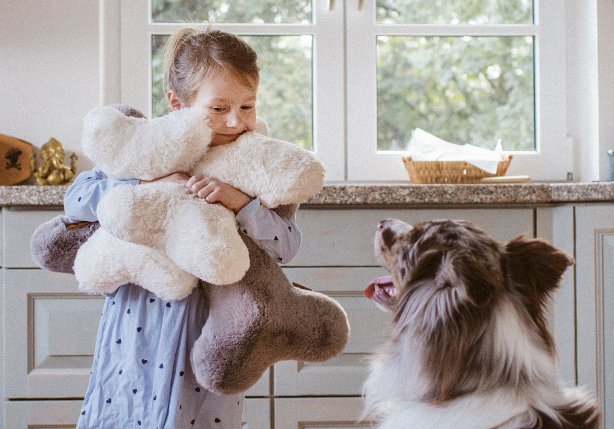 Mädchen umarmt liebevoll zwei Hyggebone Kuschelkissen in Ivory und Stone neben einem Australian Shepherd in der Küche